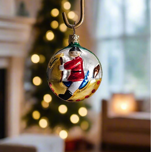 Decorative Christmas ornament with a reflection of a person in a red coat, set against a festive background with a tree and fireplace.